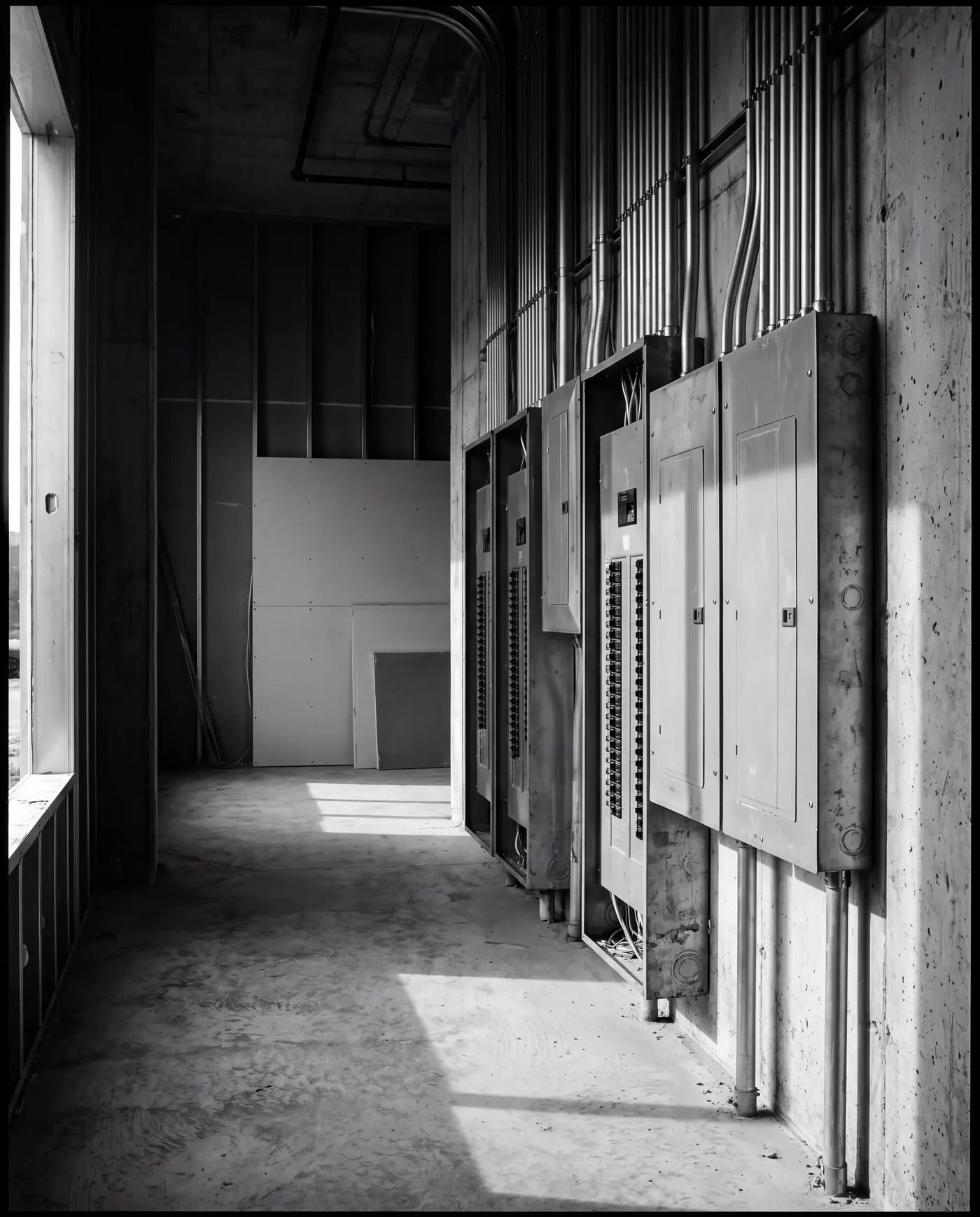 Black-and-white photograph of electrical distribution panels and conduit risers in the utility corridor of a multifamily building under construction